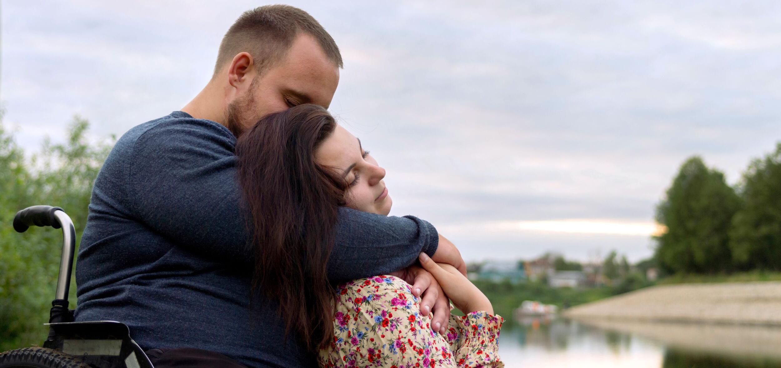 A couple sitting in front of a body of water, the man wrapping his arms loving around his partner