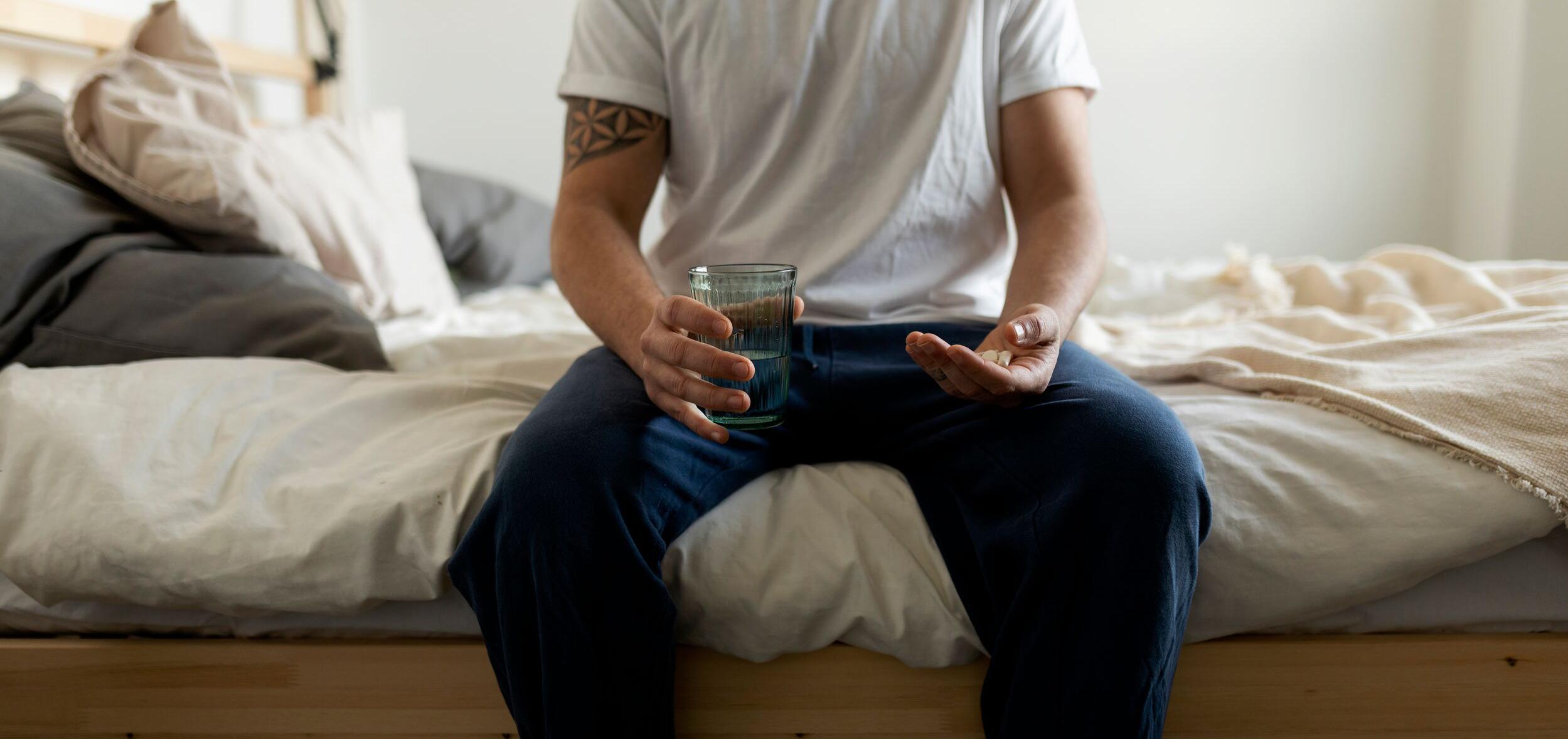 Man in jeans and a t-shirt sits on end of bed holding medications for chronic pain with MS