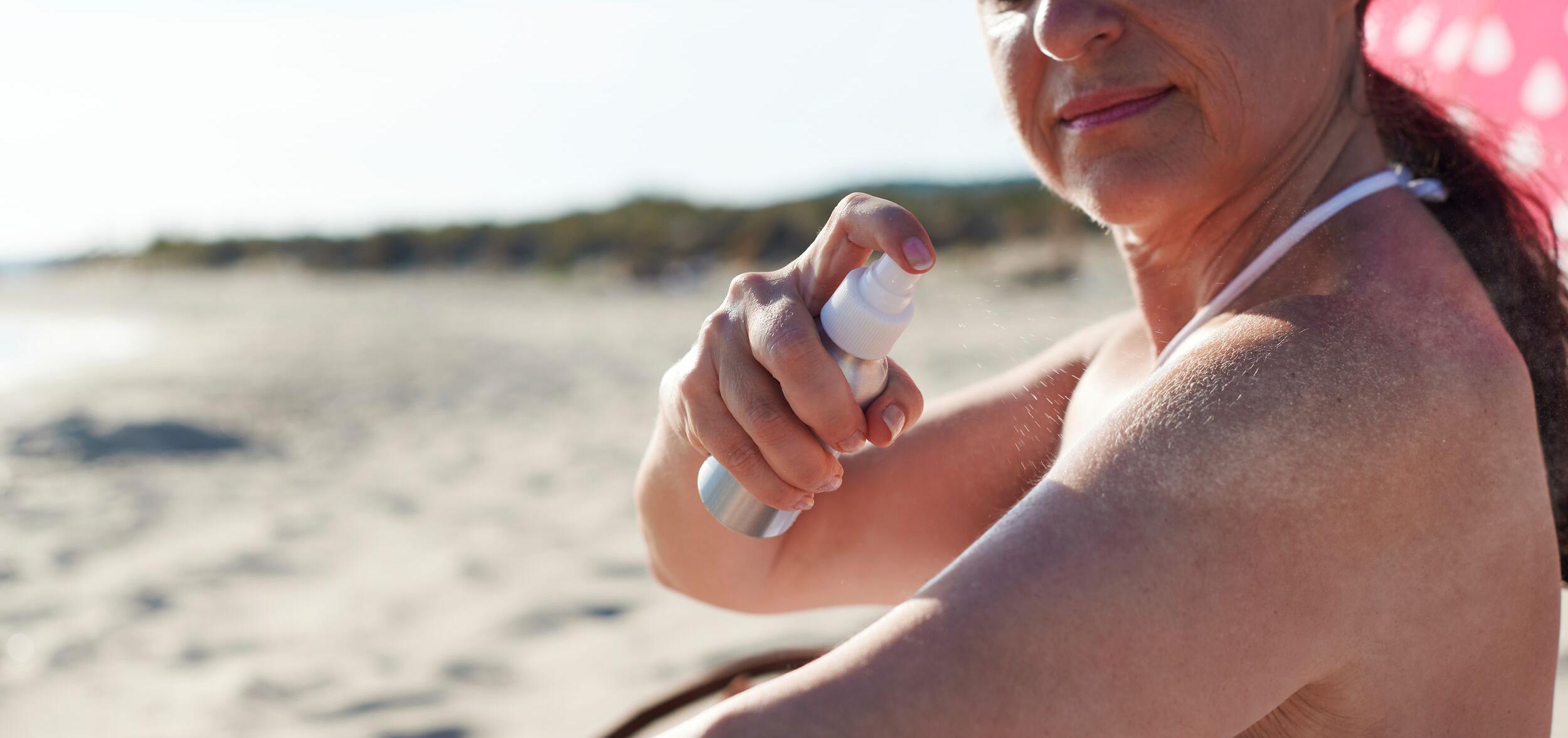 woman at the beach spraying sunscreen on her arm