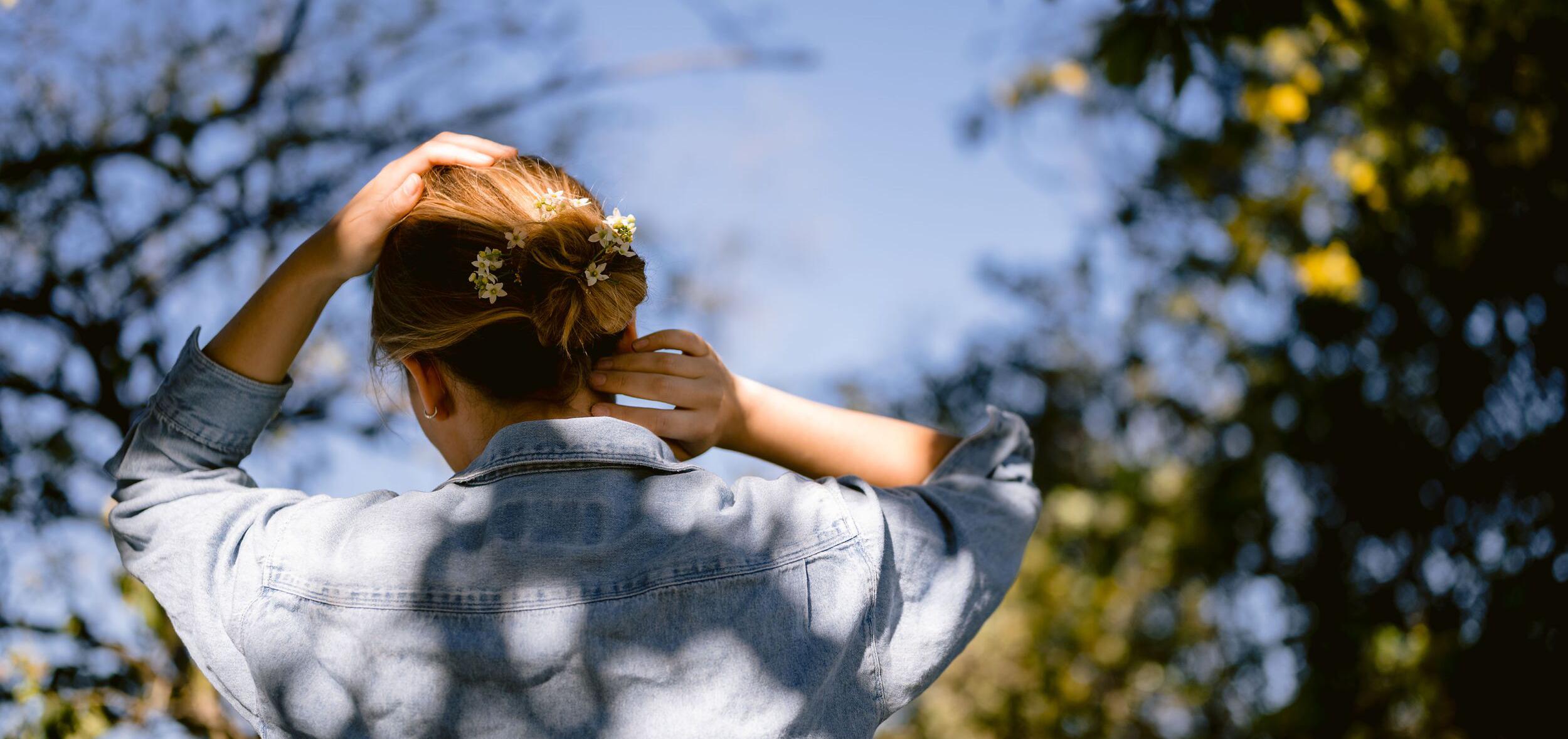 woman with bare forearms reaching toward her hair, which has flowers tucked in it