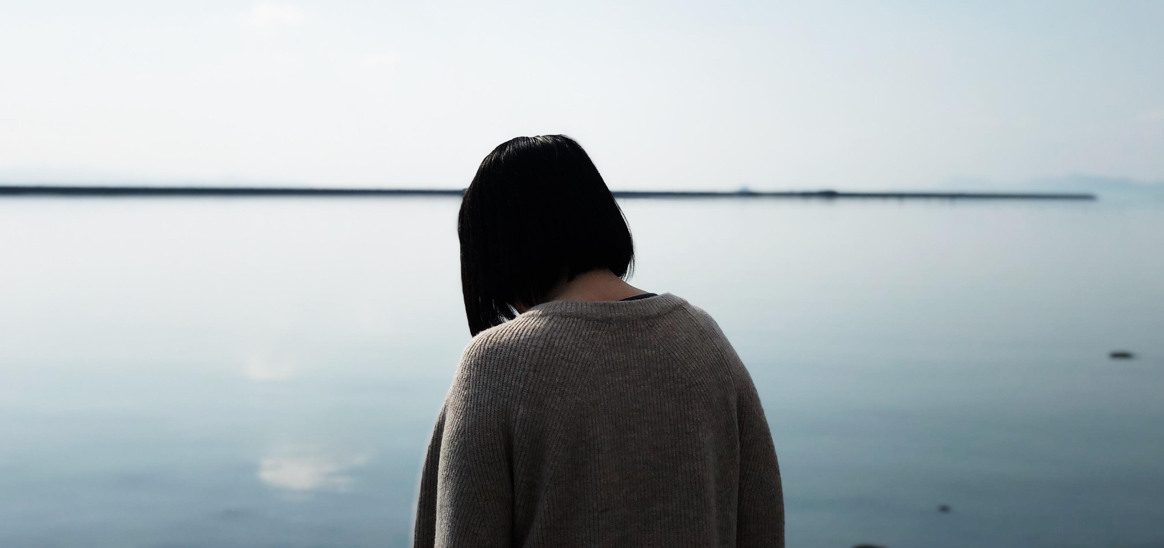 A woman with her back turned looking sadly into a body of water