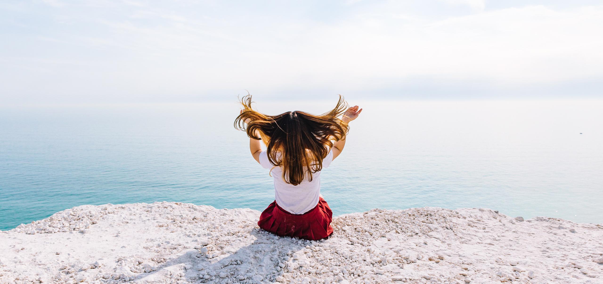 A woman sitting on a rock overlooking the ocean, her hair blowing in the wind and her arms raised