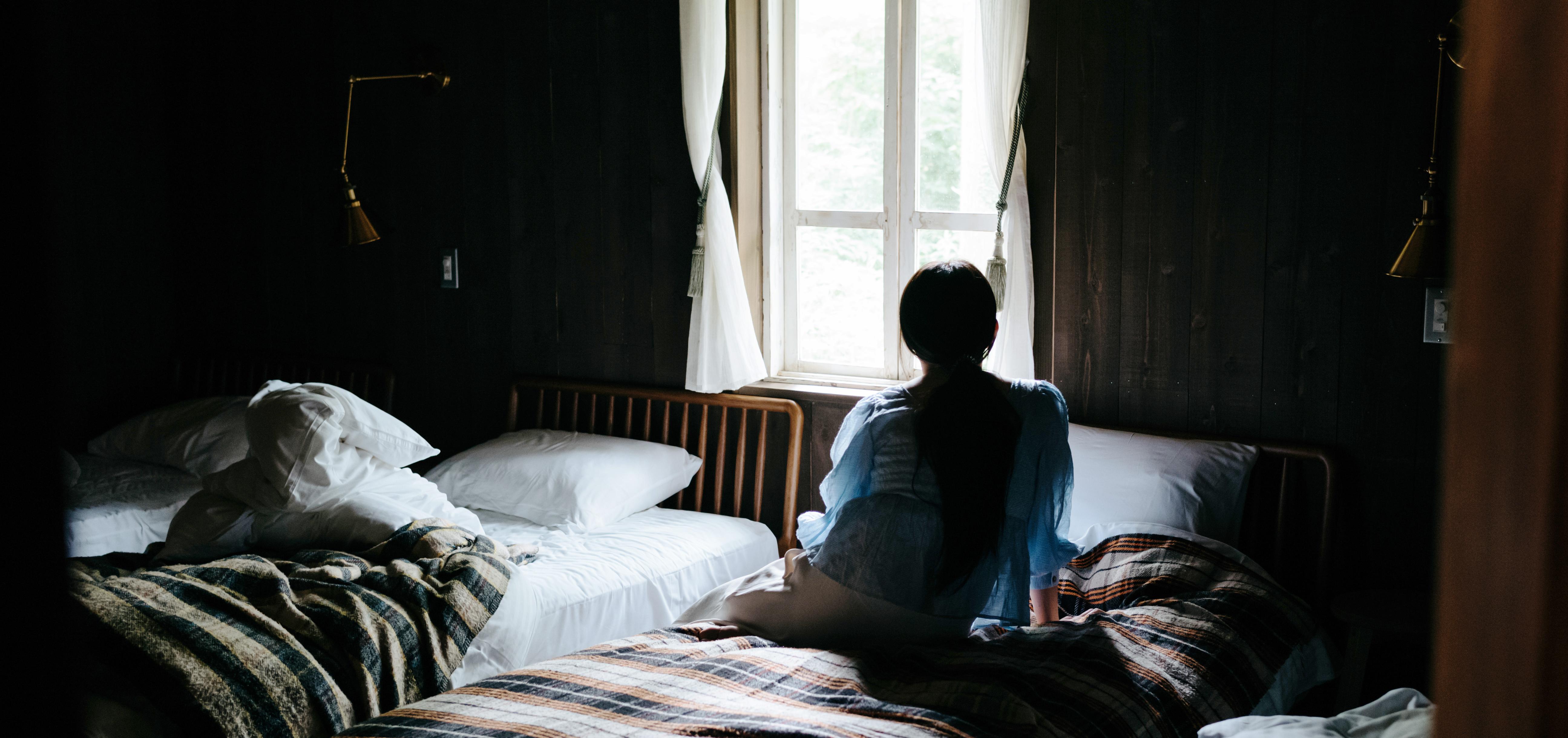 A woman in a dimly-lit bedroom sitting on a bed while looking out the window