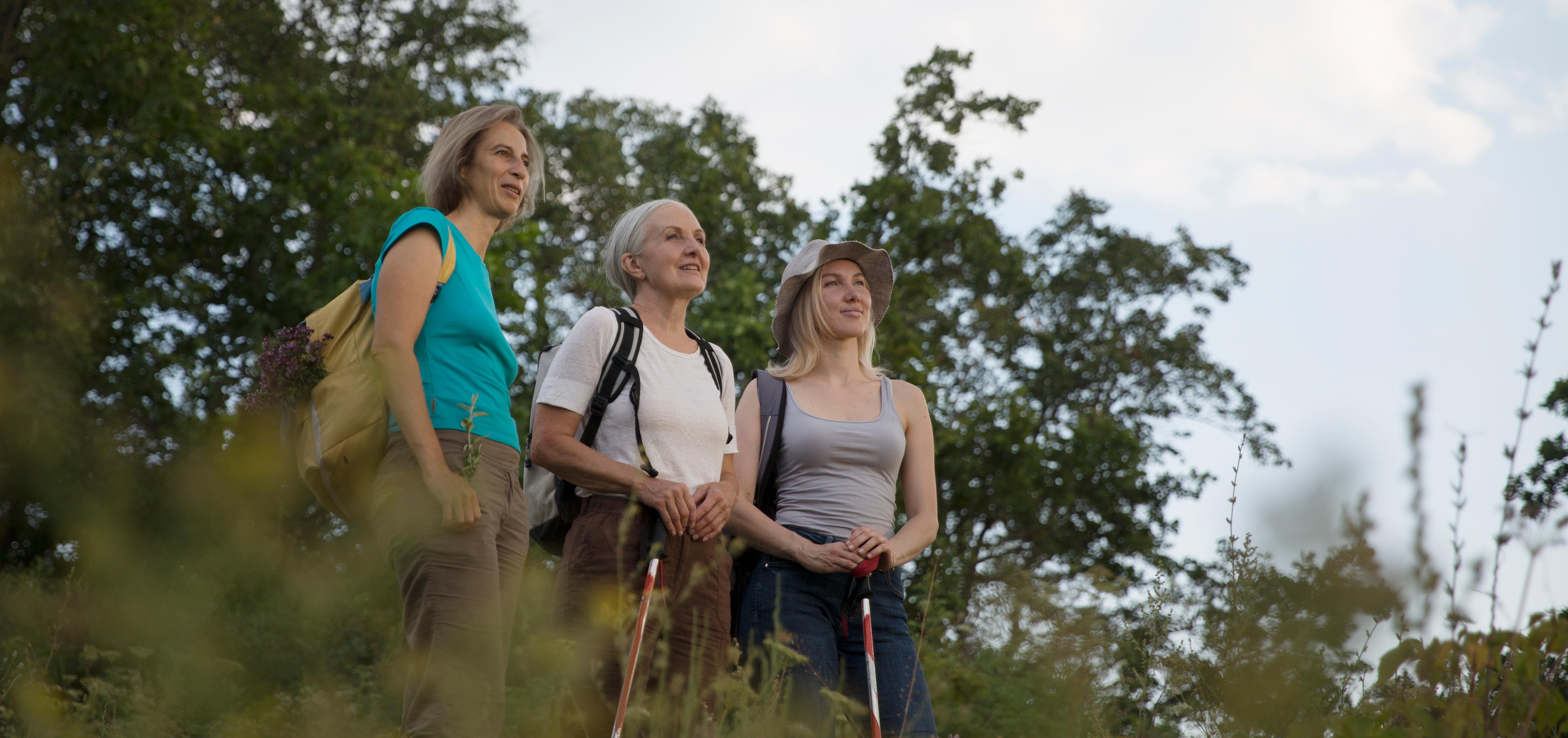 Three women, two with walking sticks, stand outside among trees