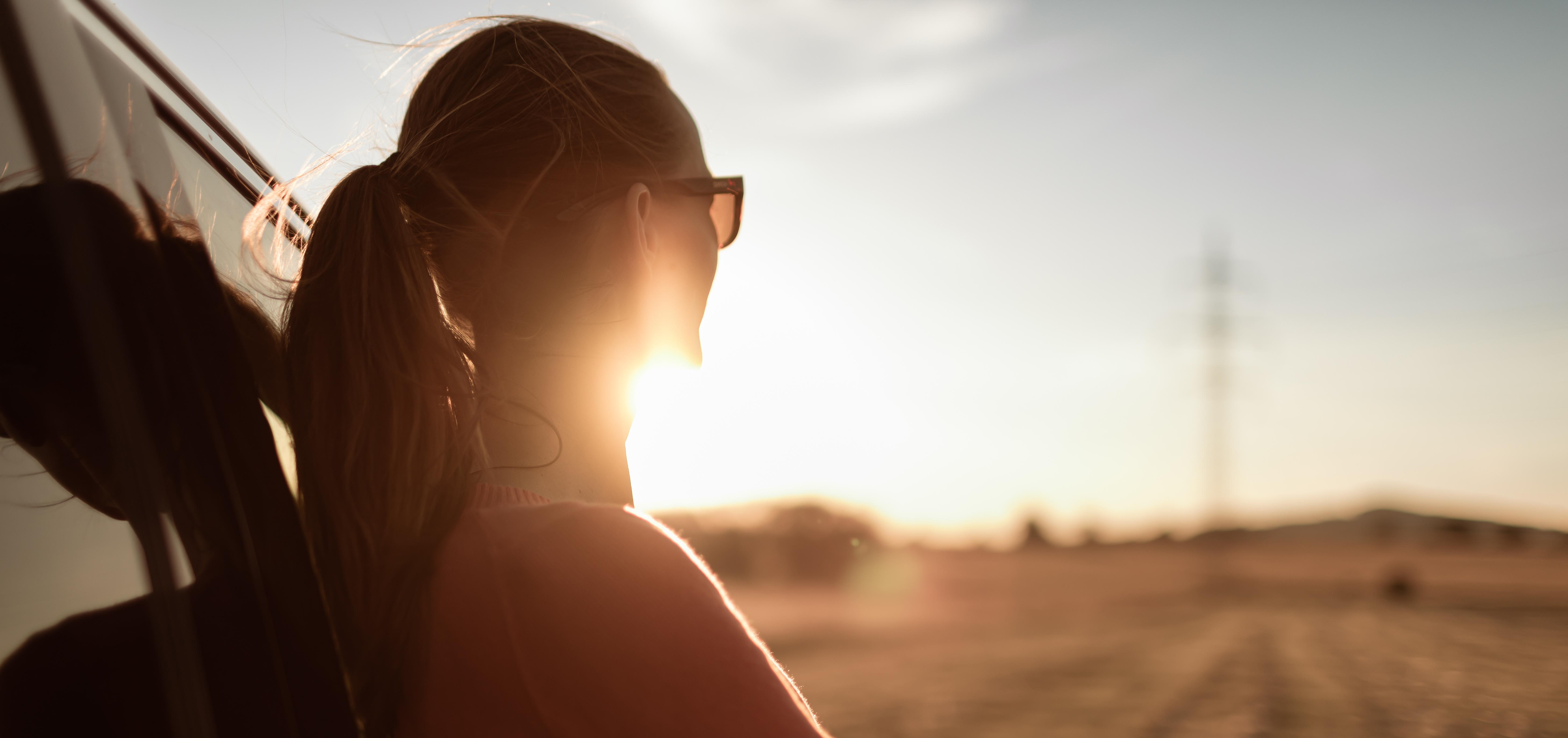 Woman leaning against a car looking calmly out toward the sun