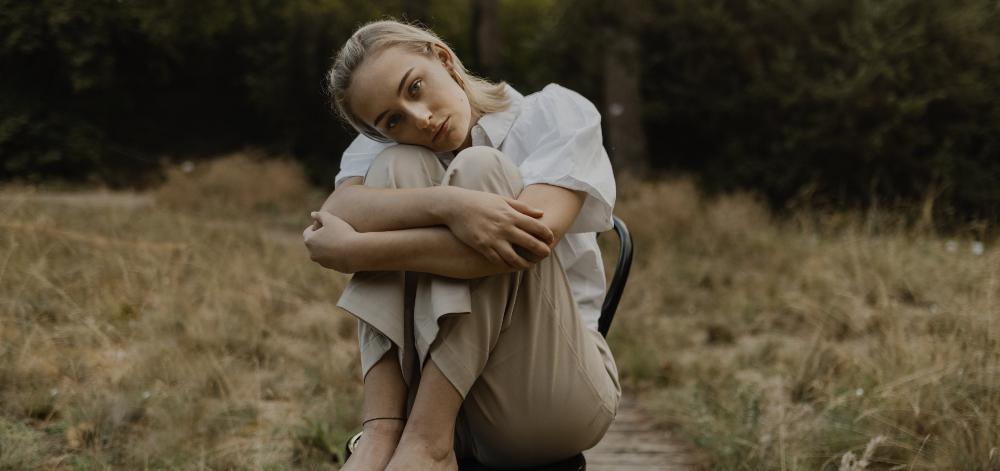 A woman in a field of grass, sitting on a chair and hugging her knees into her body, in a melancholy mood 